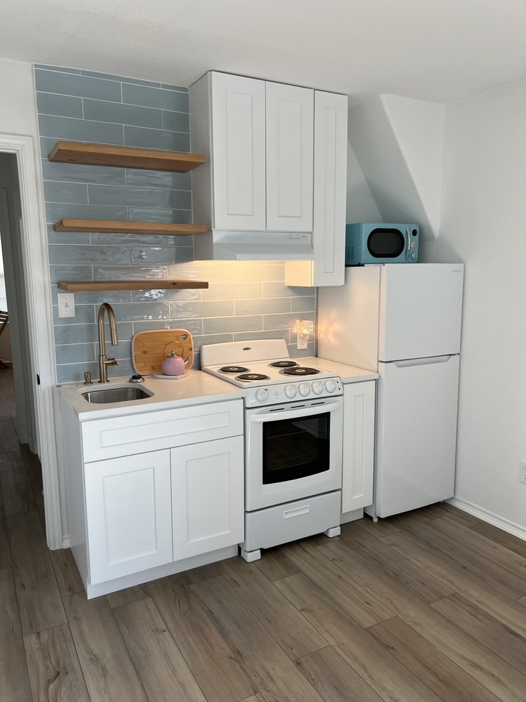 A studio kitchenette with white shaker cabinets, floating cedar shelves, blue subway tile backsplash, and white quartz countertops.